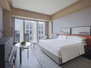 A modern hotel room with a large bed, a desk, a chair, and a view of the cityscape through the windows.