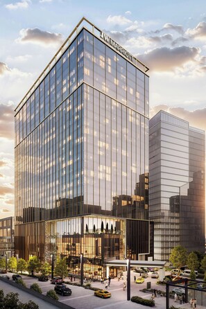 A modern hotel with a glass facade, a prominent sign, and a parking area with cars.