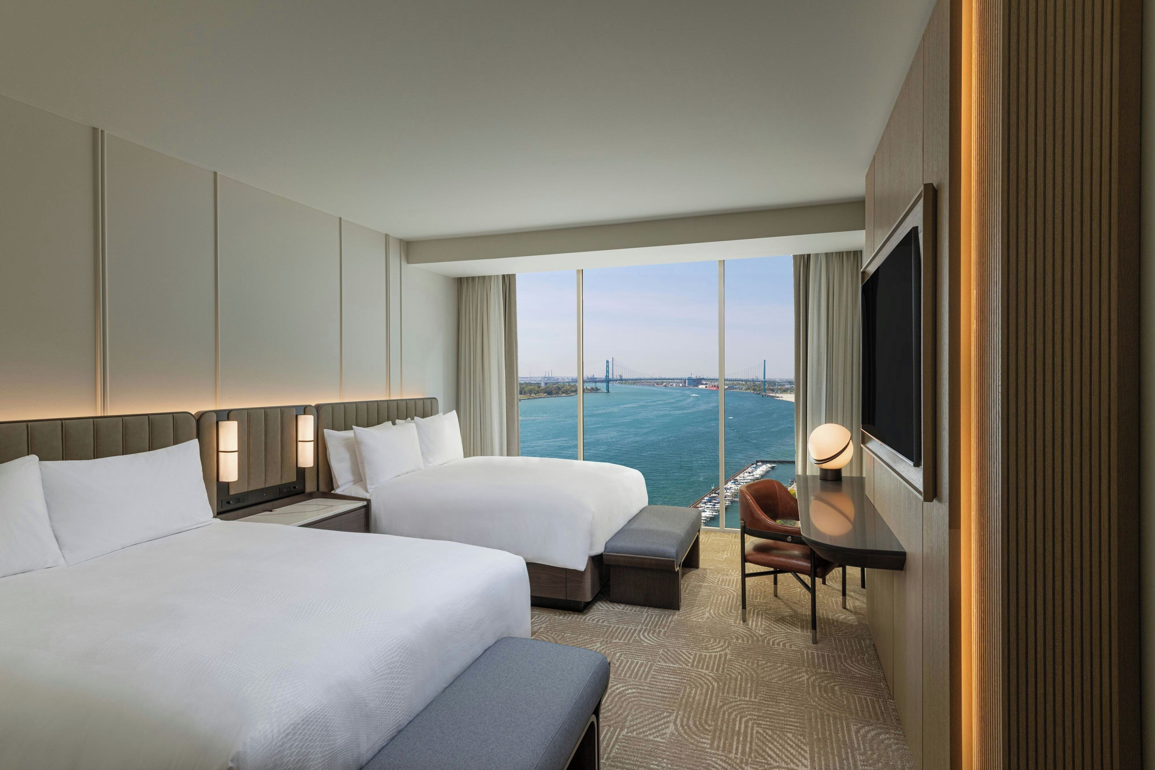 A hotel room with two beds, a desk with a chair, a TV, and a view of the sea and a bridge.