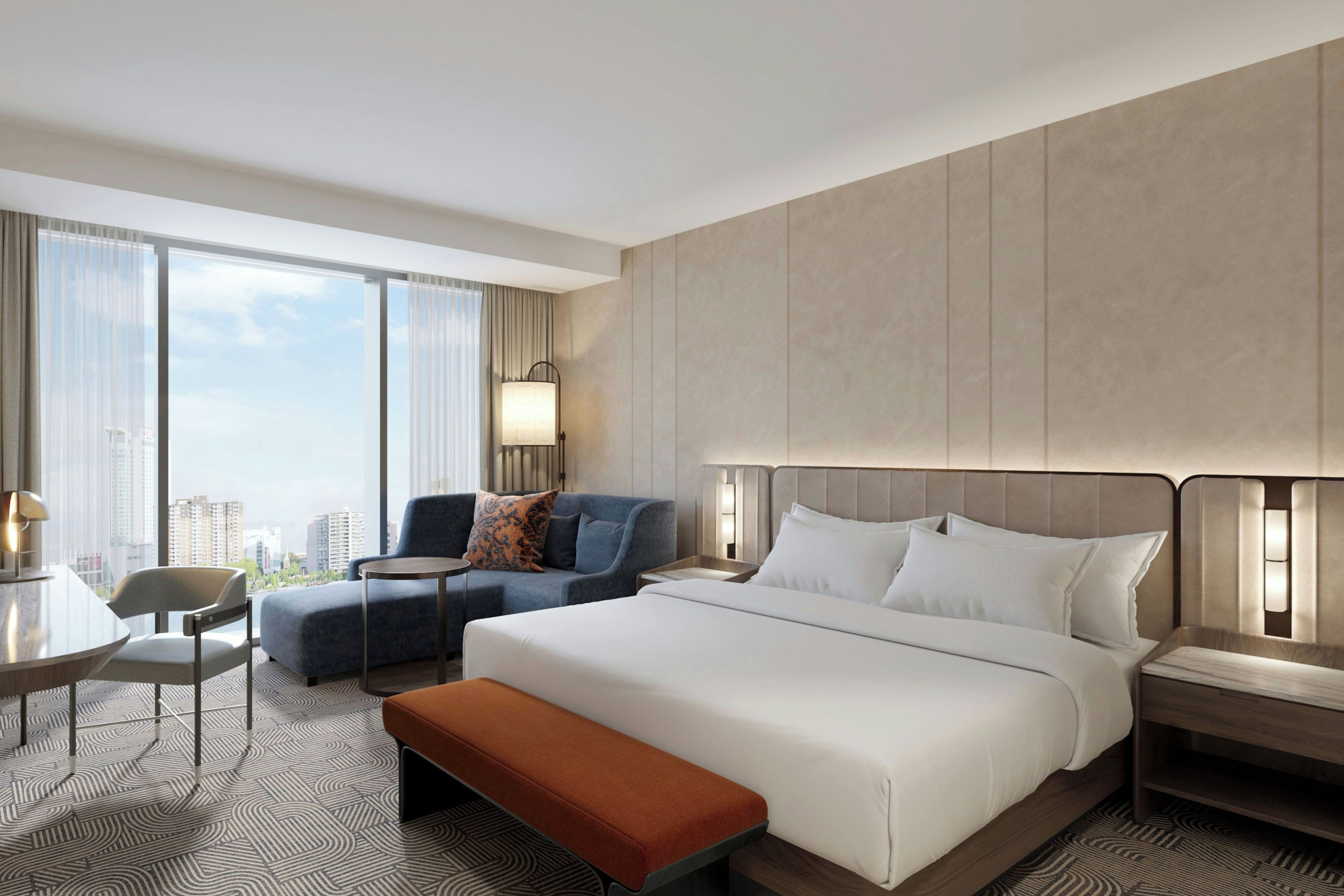 A hotel room with a large bed, a sitting area with a sofa and armchair, a small table, and a view of the city.