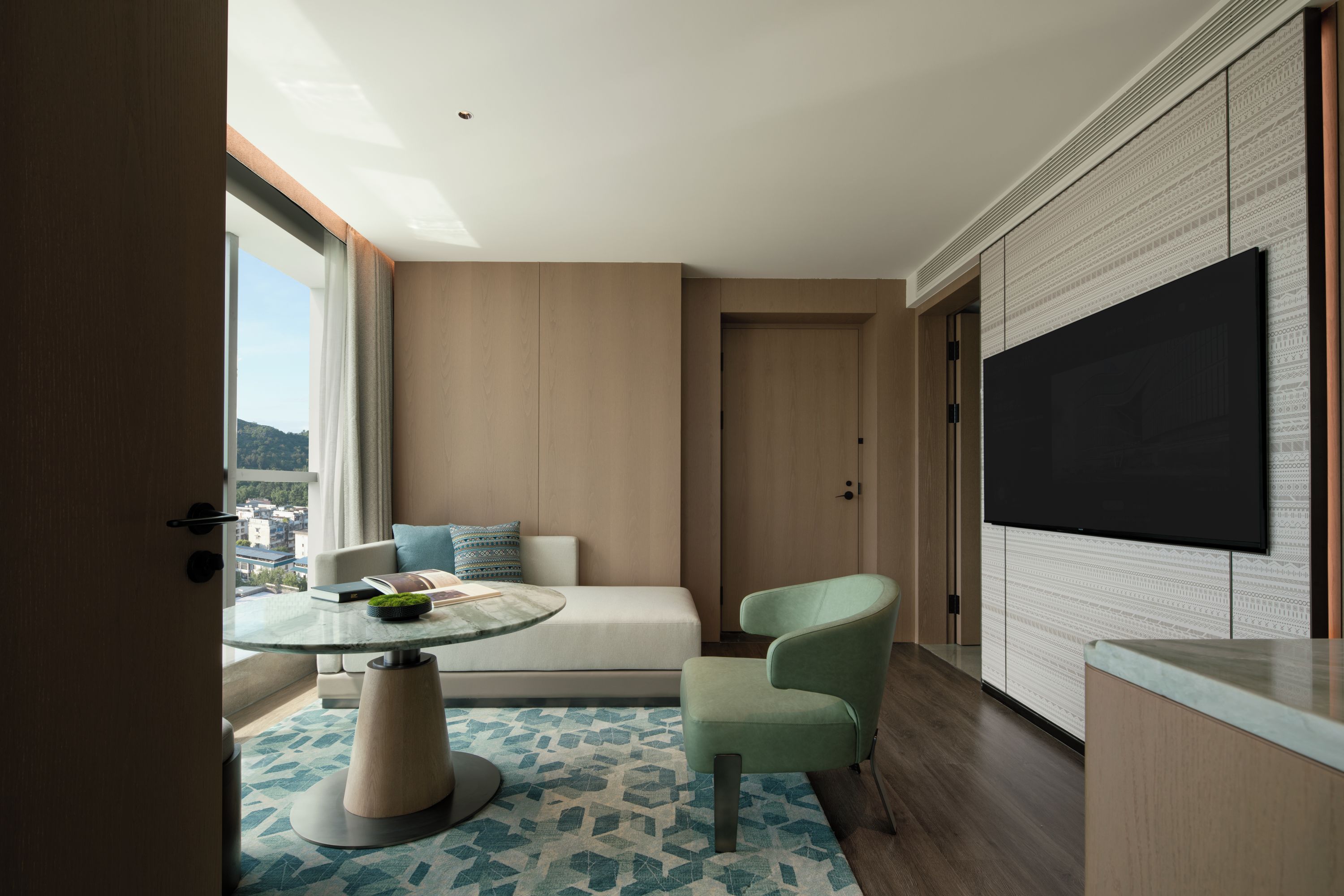 A modern hotel room with a large TV, a sofa, a round table, and a view of the city.