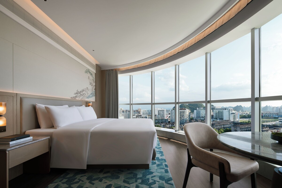 A hotel room with a large bed, a chair, a nightstand, and a view of the city through large windows.