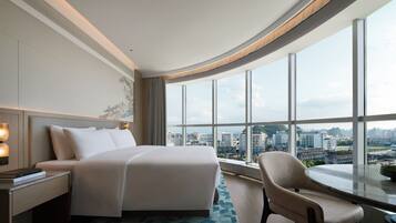 Habitación de hotel con una cama grande, una silla, una mesita de noche y vistas de la ciudad a través de grandes ventanales.