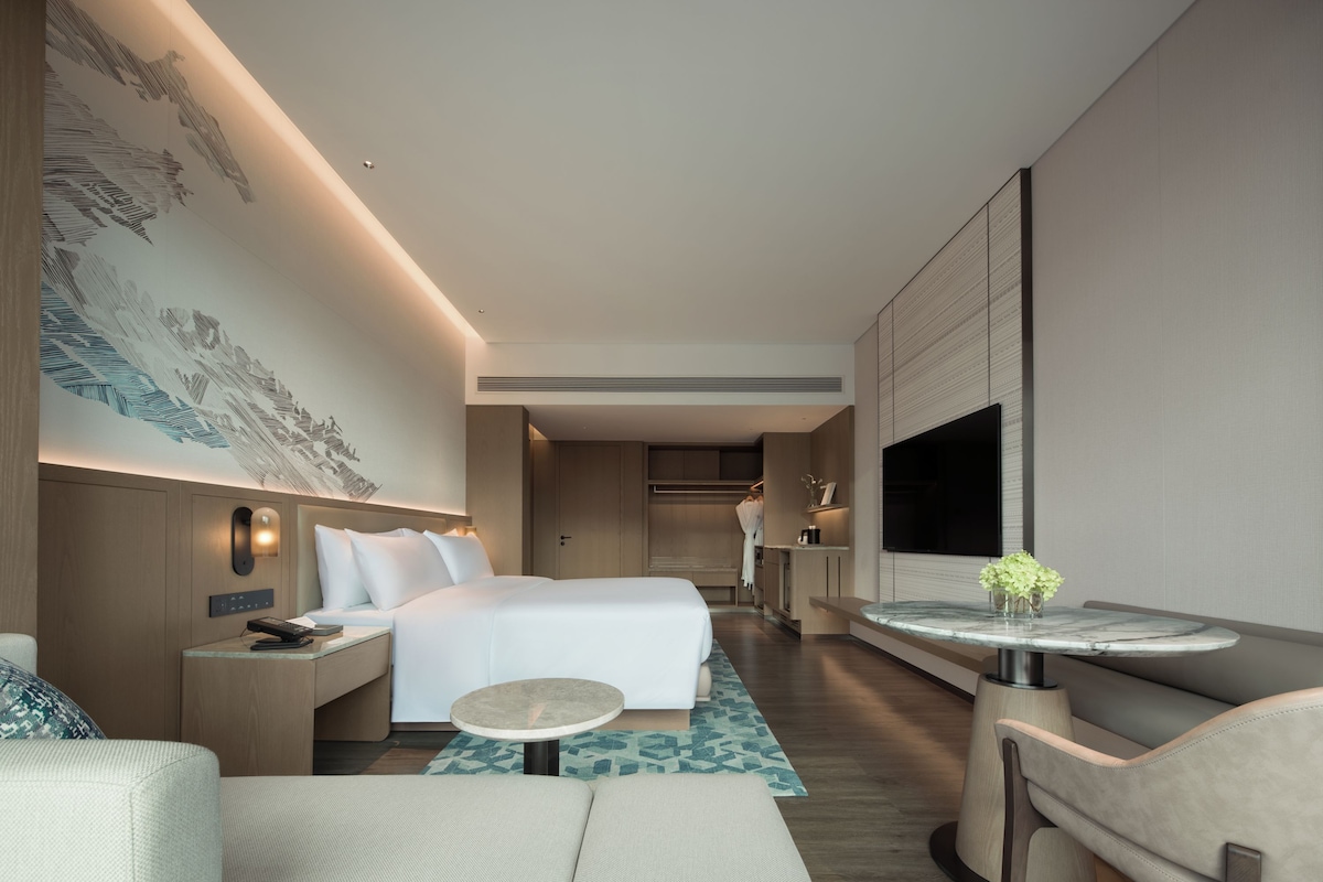 A modern hotel room with a bed, bedside table, lamp, and a small round table with a vase of flowers.