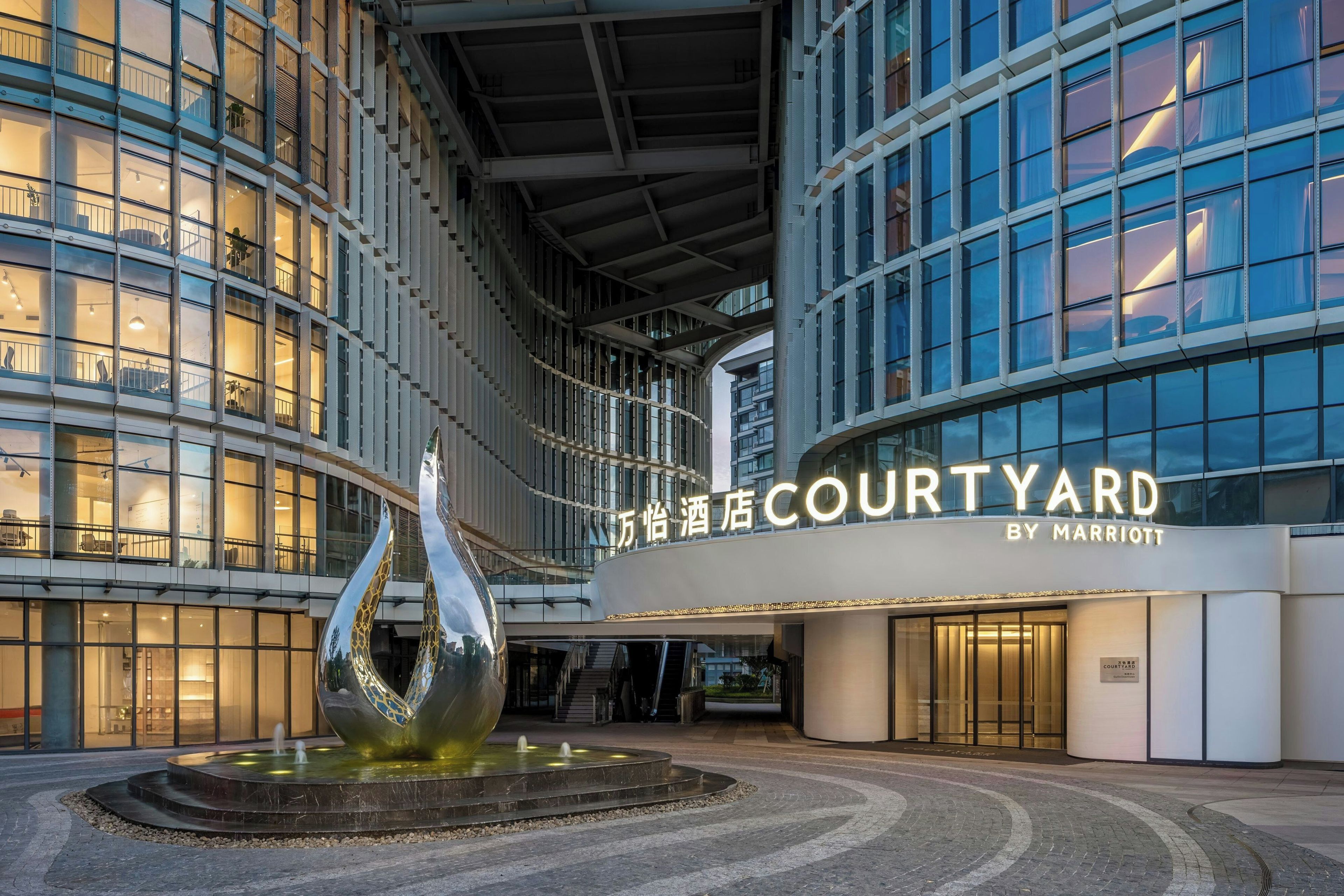 Entrada de un hotel Courtyard by Marriott con fachada de vidrio moderna, una fuente central y un área de acceso bien iluminada.