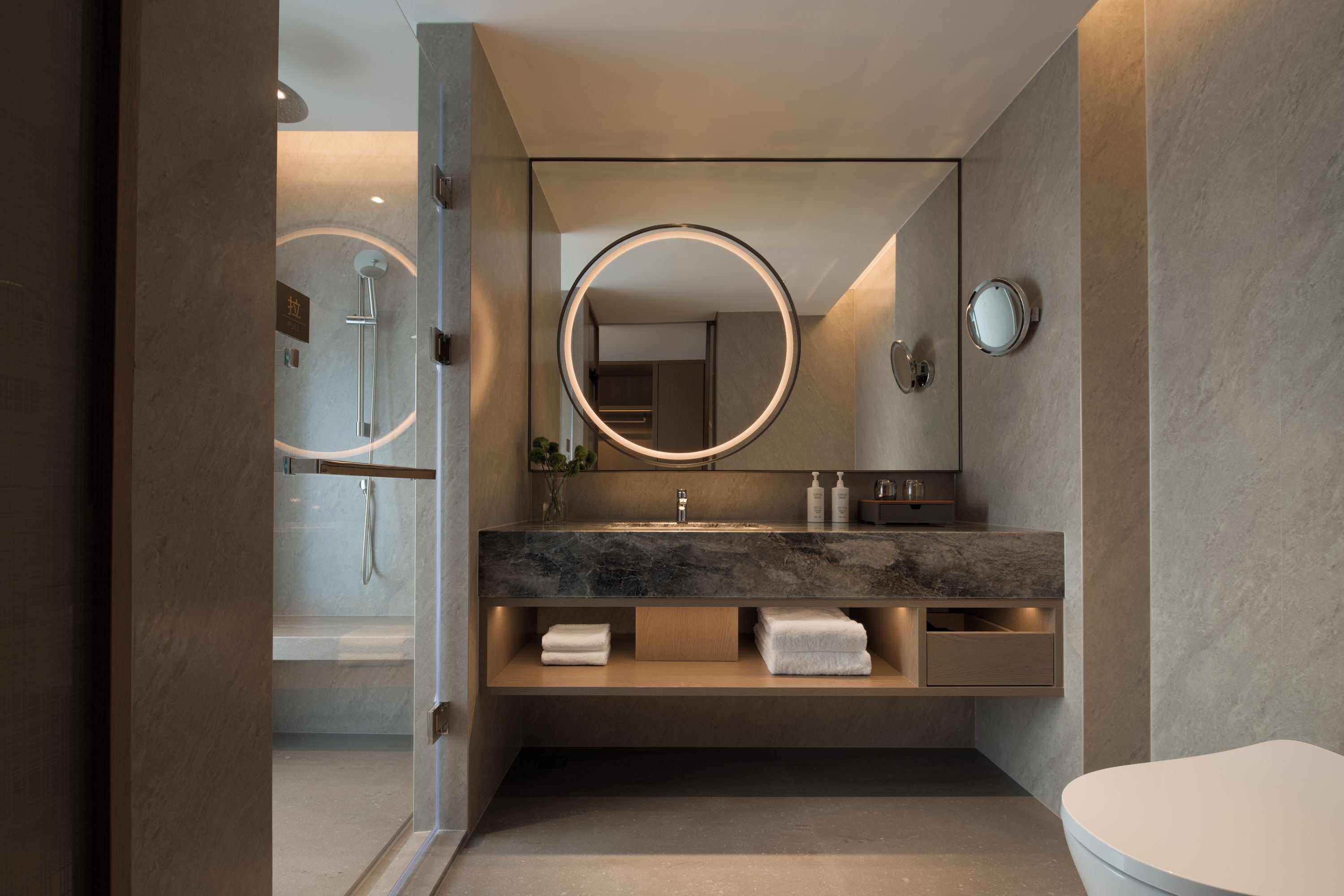 Un baño moderno con ducha envuelta en vidrio, tocador de mármol y un espejo redondo.