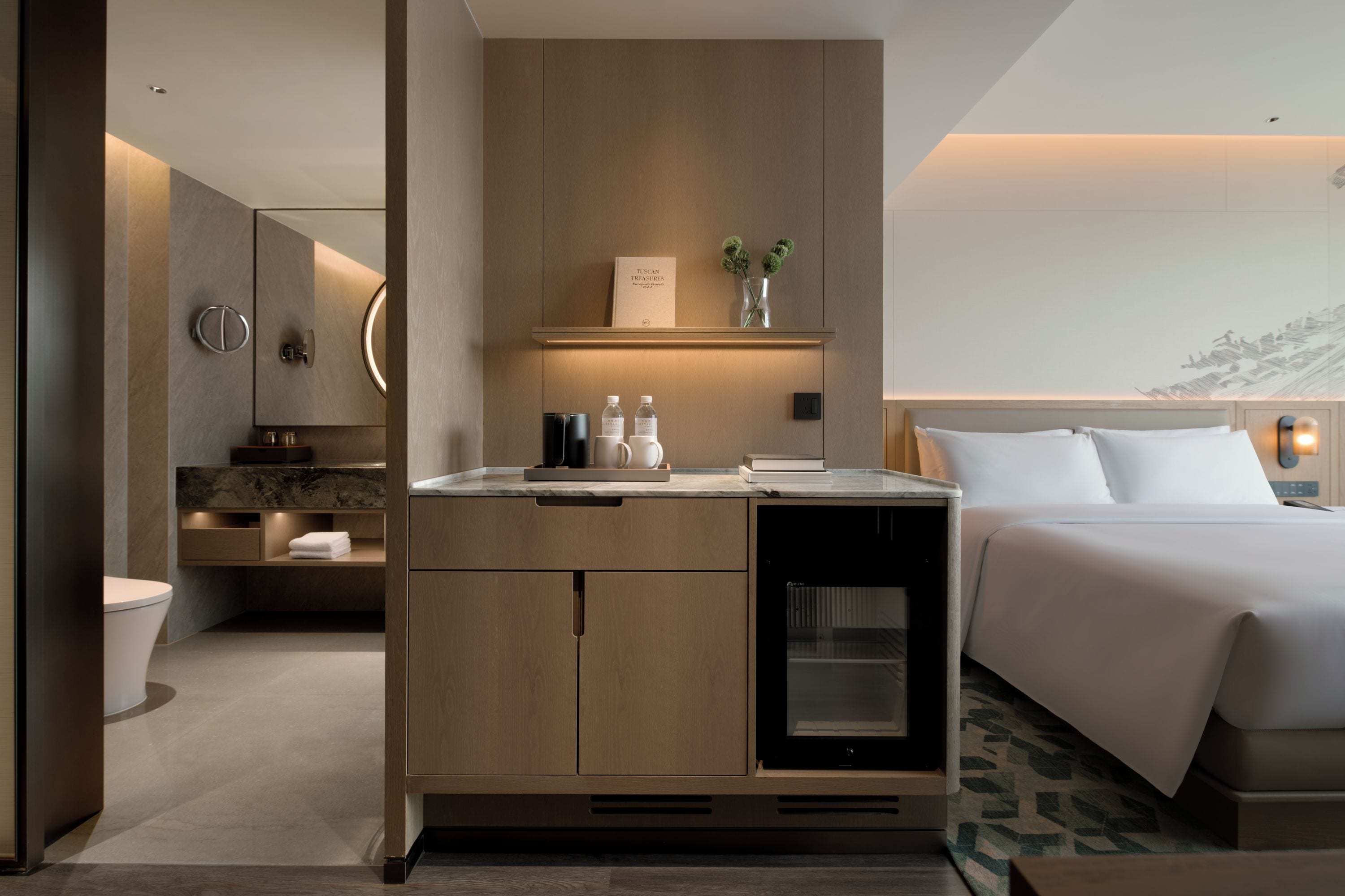 A modern hotel room with a bed, a minibar, a bathroom, and a view of a mountain.