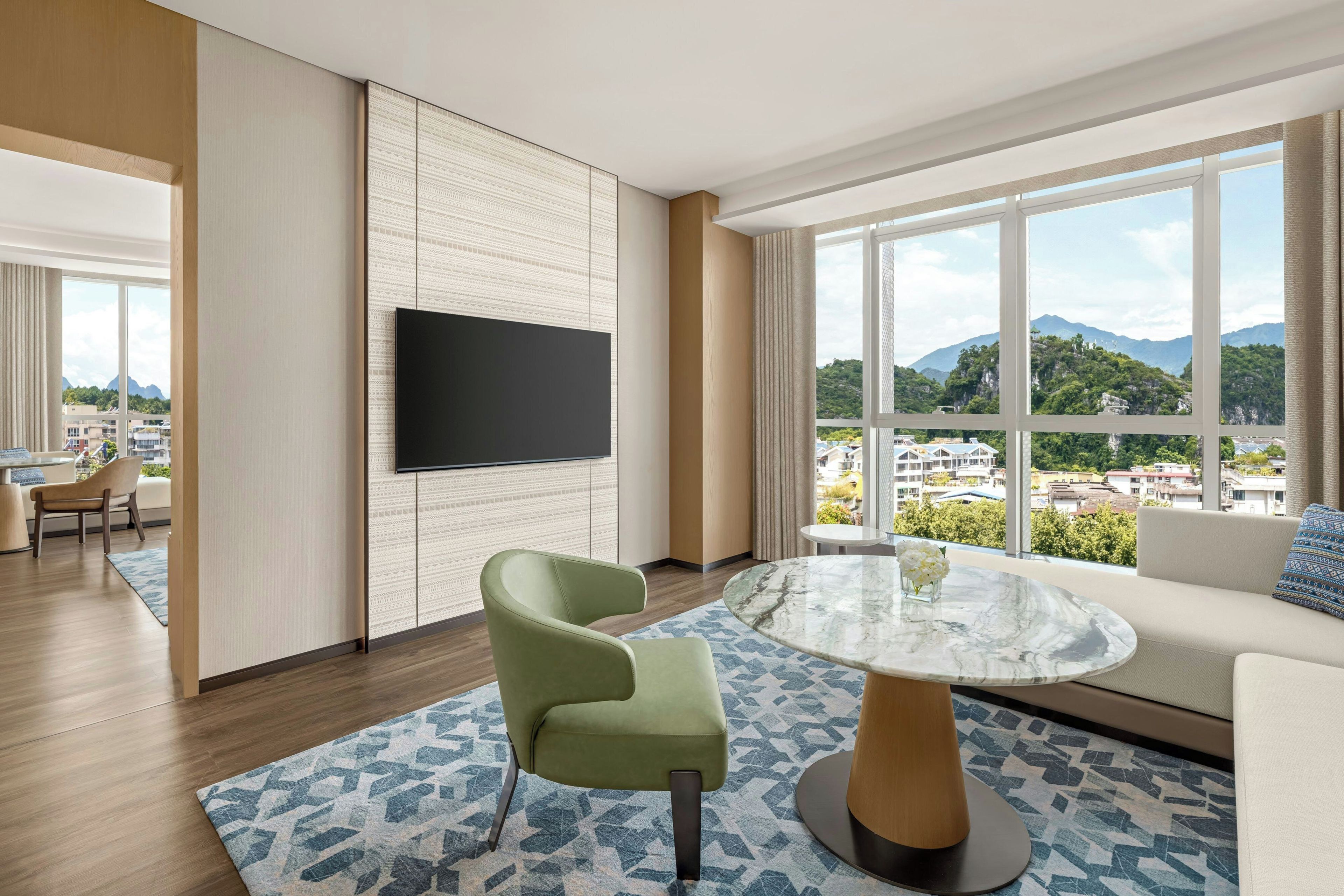 A modern hotel room with a large window, a flat-screen TV, a green armchair, a round glass table, and a patterned rug.