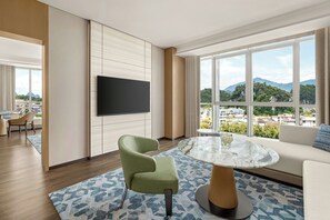 Ein modernes Hotelzimmer mit großem Fenster, einem Flachbildfernseher, einem grünen Sessel, einem runden Glastisch und einem gemusterten Teppich.