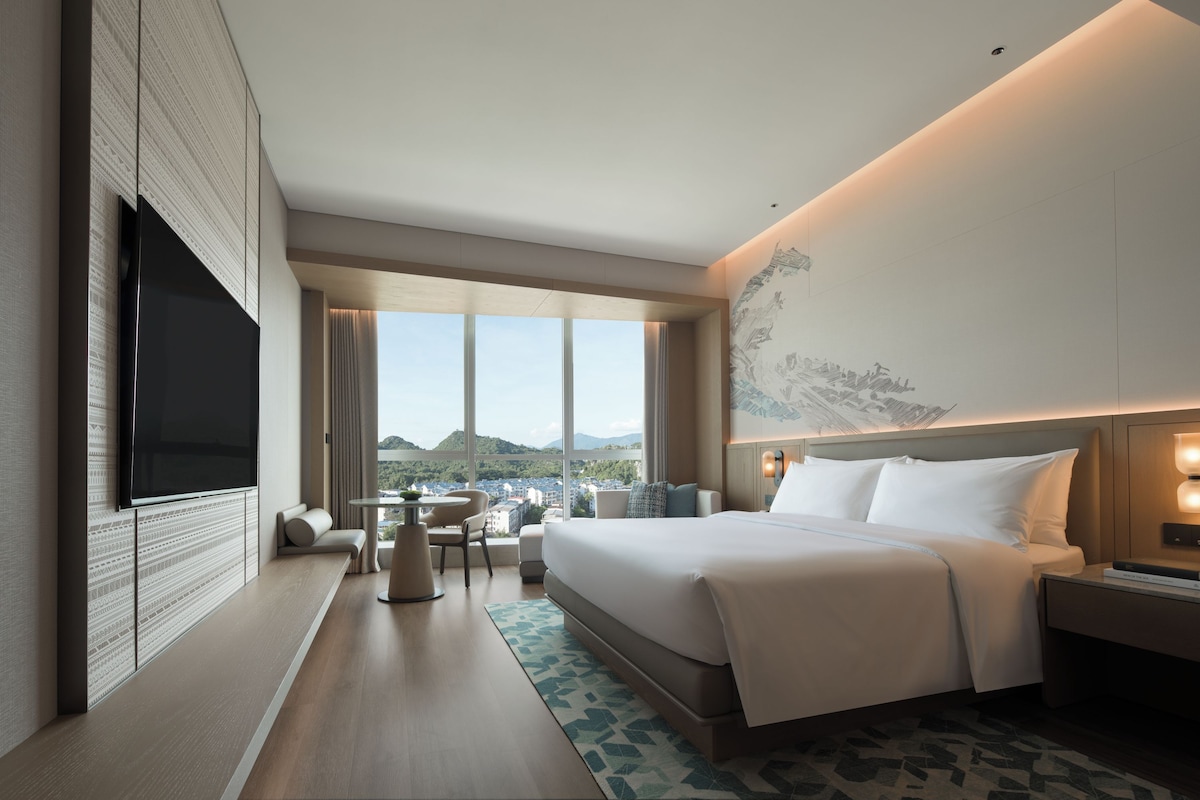 A modern hotel room with a large bed, a flat-screen TV, a dining area, and a view of the cityscape.