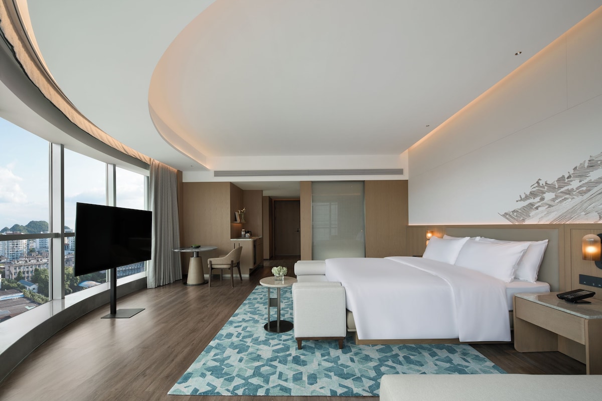 A modern hotel room with a large bed, a flat-screen TV, a desk, and a view of the city.