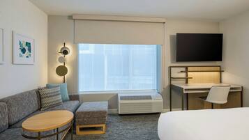 A hotel room with a bed, a desk, a TV, a sofa, a small table, and a window with blinds.
