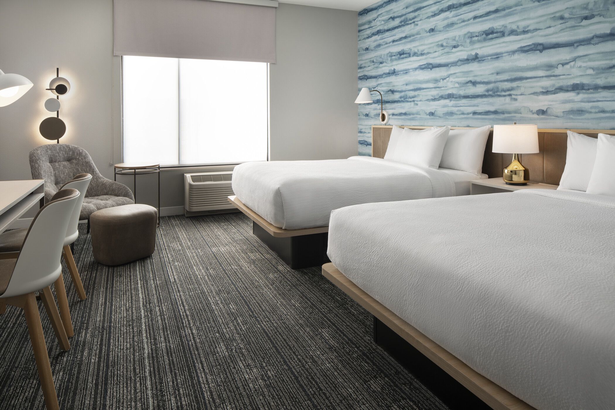 A hotel room with two beds, a desk, a chair, an air conditioning unit, and a wall with a blue abstract pattern.