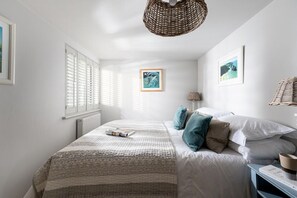 A bedroom with a large bed, a bedside table, a lamp, and a wicker chair. - 3 Bed in Mawgan Porth (oc-b584383) (Mawgan Porth)