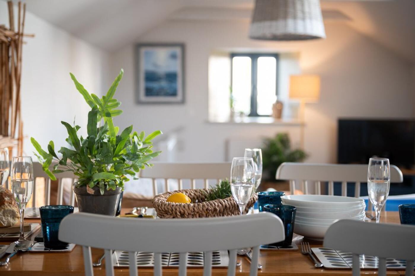 Mesa de comedor dispuesta con sillas blancas, una planta en maceta, una cesta tejida con un limón y varios vasos de agua.