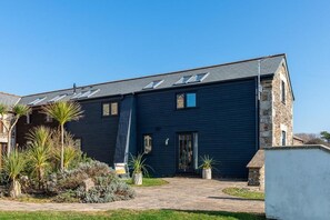 A two-story building with a dark exterior, a stone tower, and a paved driveway with landscaping. - 3 Bed in Mawgan Porth (oc-b584383) (Mawgan Porth)
