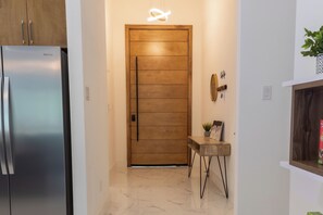 Un couloir avec une porte en bois, un réfrigérateur, une petite table avec une plante et un luminaire fixé au mur.