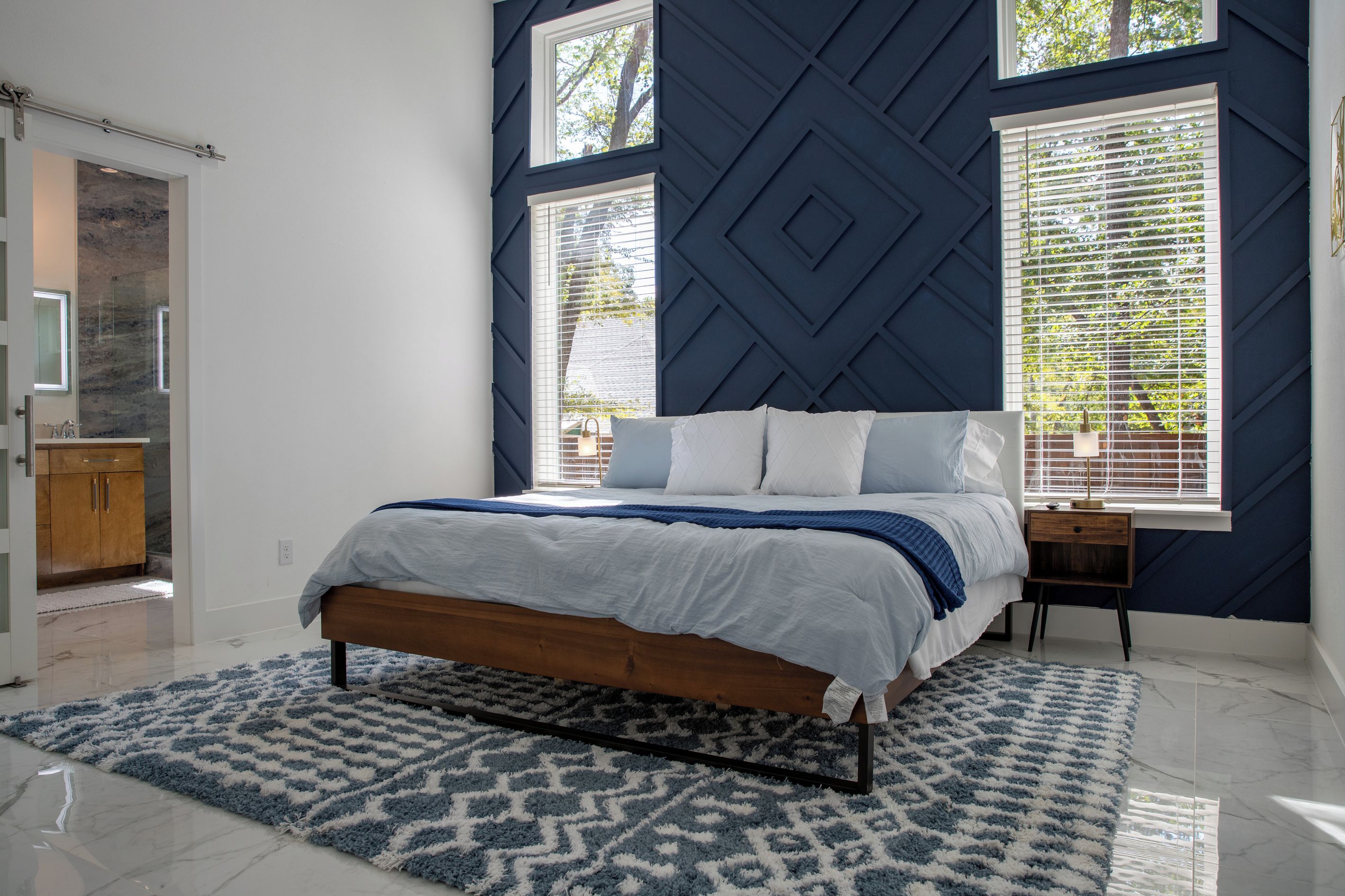 Un dormitorio con cama, mesita de noche, ventana con persianas y puerta que da a otra habitación.