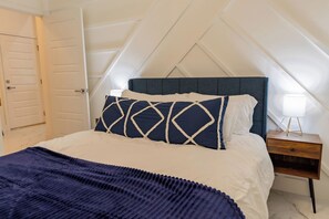 Ein Schlafzimmer mit schrägen Wänden, einem Bett mit einem blau-weißen Kissenmuster, einem Nachttisch mit einer Lampe und einem Holznachtkästchen.