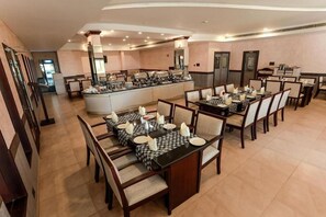 A hotel dining area with multiple tables and chairs, a buffet station, and a kitchen in the background. - Hotel Bluewaters Cherai (Kochi)
