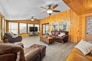 Ein geräumiges Wohnzimmer mit einem braunen Ledersofa, einer braunen Lederliege und einem Couchtisch aus Holz. Eine Deckenlampe und ein Fernseher auf einer Holzkommode sind ebenfalls vorhanden.