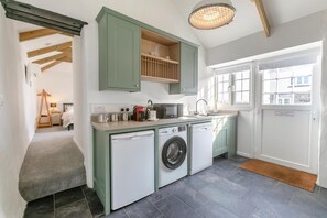 A compact kitchen with a washing machine, a microwave, and a sink. There is a wooden shelf above the sink and a door with a sign. - 1 Bed in Summercourt (oc-o36891) (Summercourt)