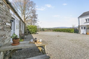 A gravel courtyard with a wooden table set for breakfast, a stone building, and a hedge in the background. - 1 Bed in Summercourt (oc-o36891) (Summercourt)