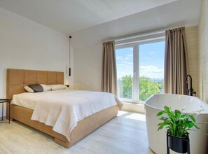 A bedroom with a bed, a window, a view of greenery, a bathtub, and a potted plant.