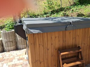 A hot tub with a wooden exterior and a grey cover, situated outdoors with a brick pathway and greenery in the background.