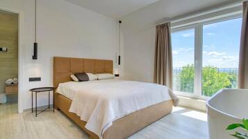 A modern bedroom with a large bed, a bedside table, a hanging light, and a view of greenery through a window.