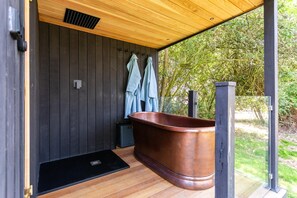 Eine Holzterrasse mit einer großen Kupferbadewanne, einem schwarzen Heizkörper und einem Handtuchhalter mit Handtüchern.