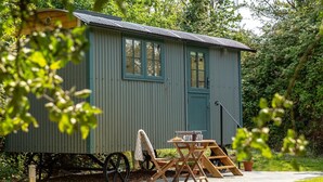 A small, single-story building with a corrugated metal exterior, a door, and windows. There is a table with chairs and a bench outside. - 1 Bed in West Yelland (oc-o45728) (Braunton)