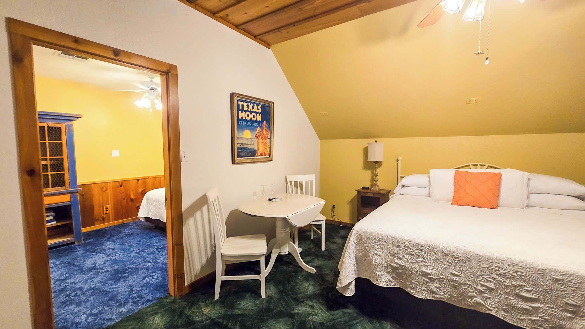 Ein Zimmer mit einem Bett, einem Tisch, einem Stuhl, einem Plakat und einer Tür zu einem anderen Raum.