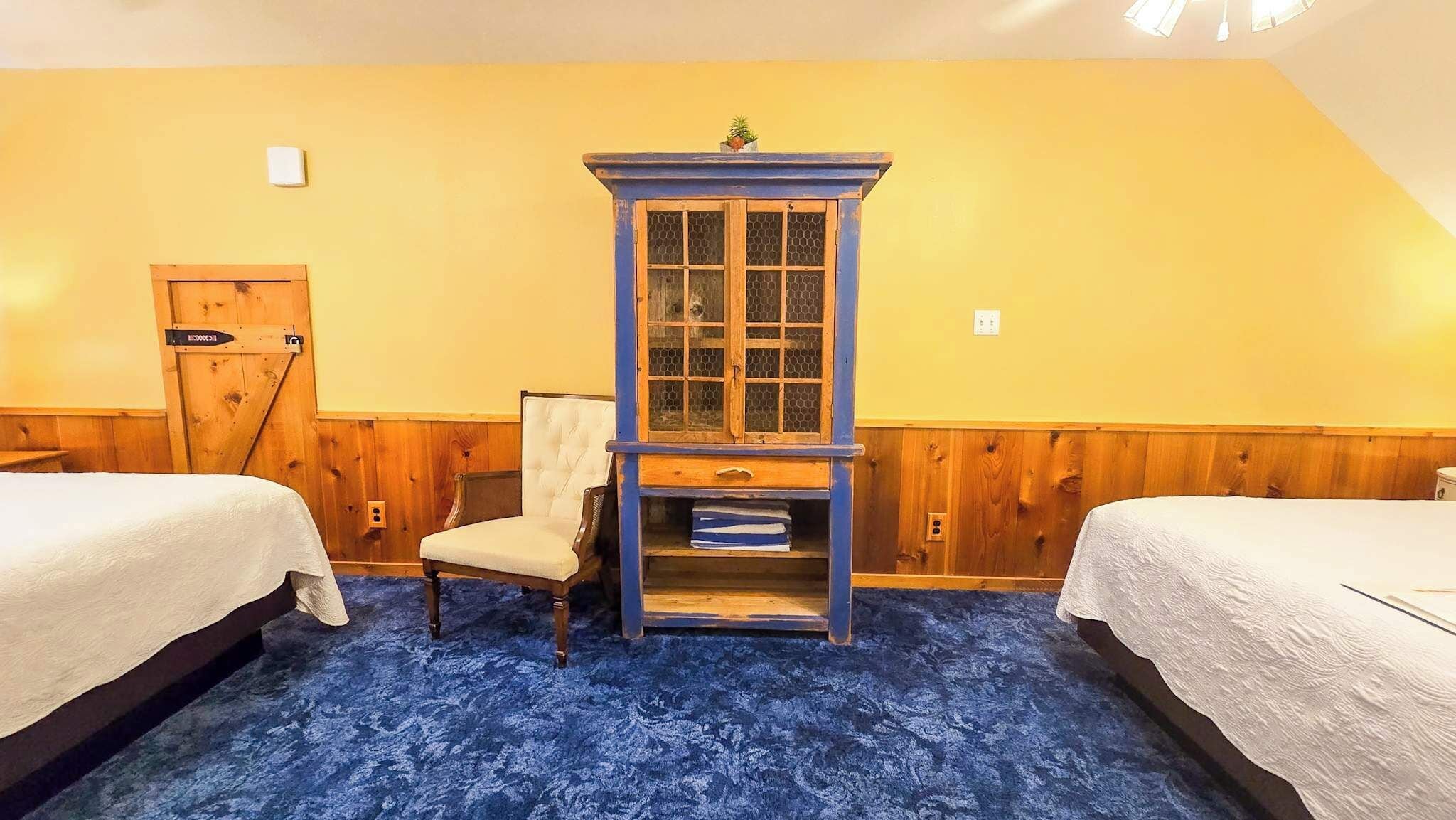 Una habitación con dos camas, un armario azul con puertas de vidrio, una silla y una puerta de madera.