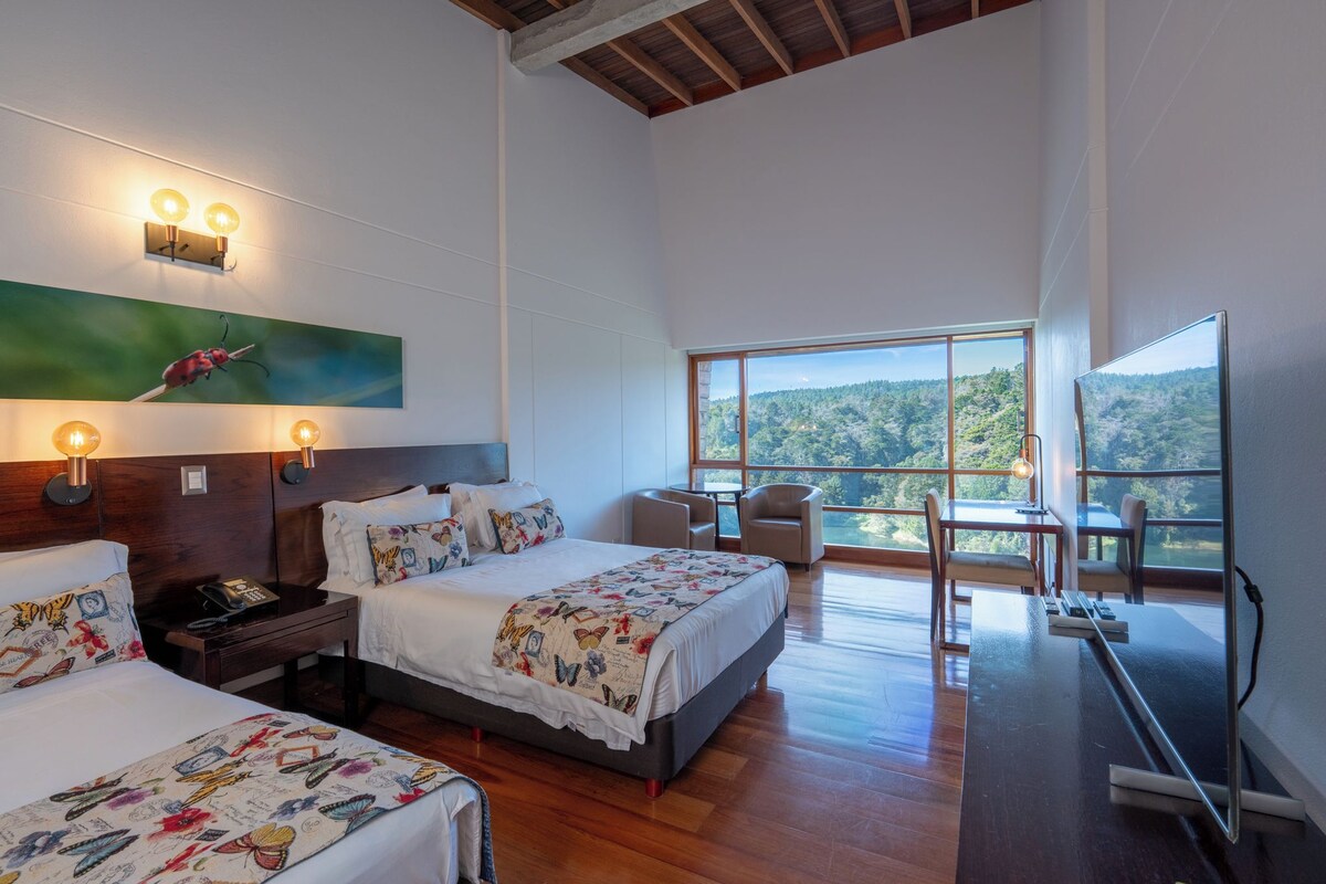 A hotel room with two beds, a desk, a chair, and a large window offering a forest view.