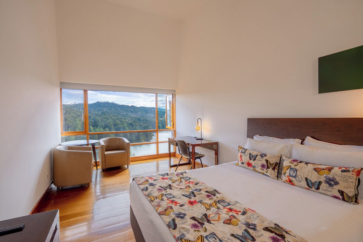 A hotel room with a large bed, a desk, a chair, and a view of a forest through a large window.
