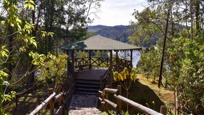 Um gazebo de madeira com telhado, rodeado por árvores e um caminho de pedra que o leva.