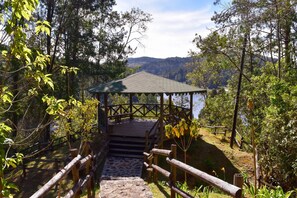 Un pabellón de madera con techo, rodeado de árboles y un sendero de piedra que conduce a él.