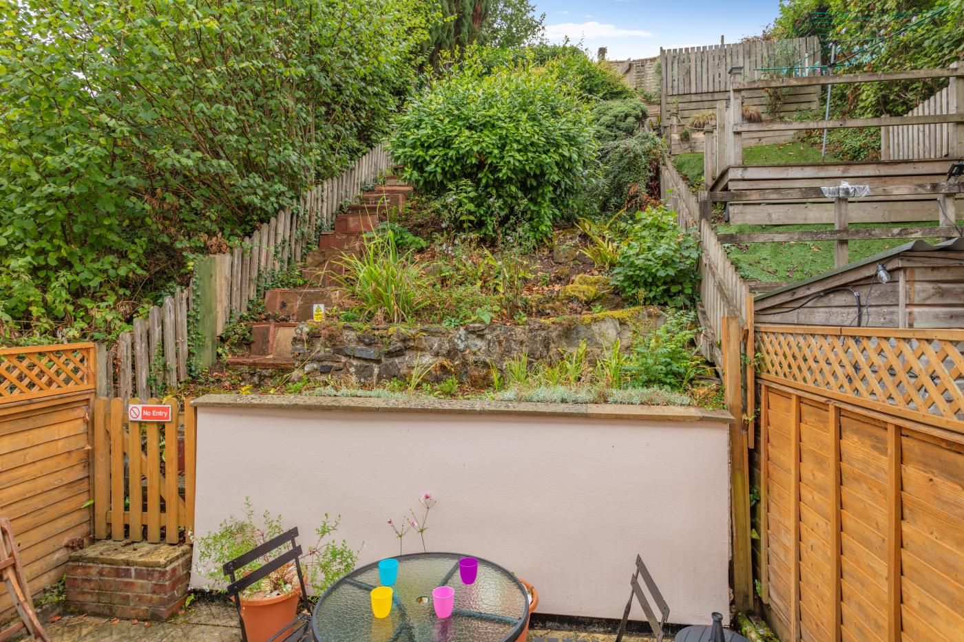 A backyard with a wooden fence, a 'No Entry' sign, a glass table with colorful cups, and a stone wall.