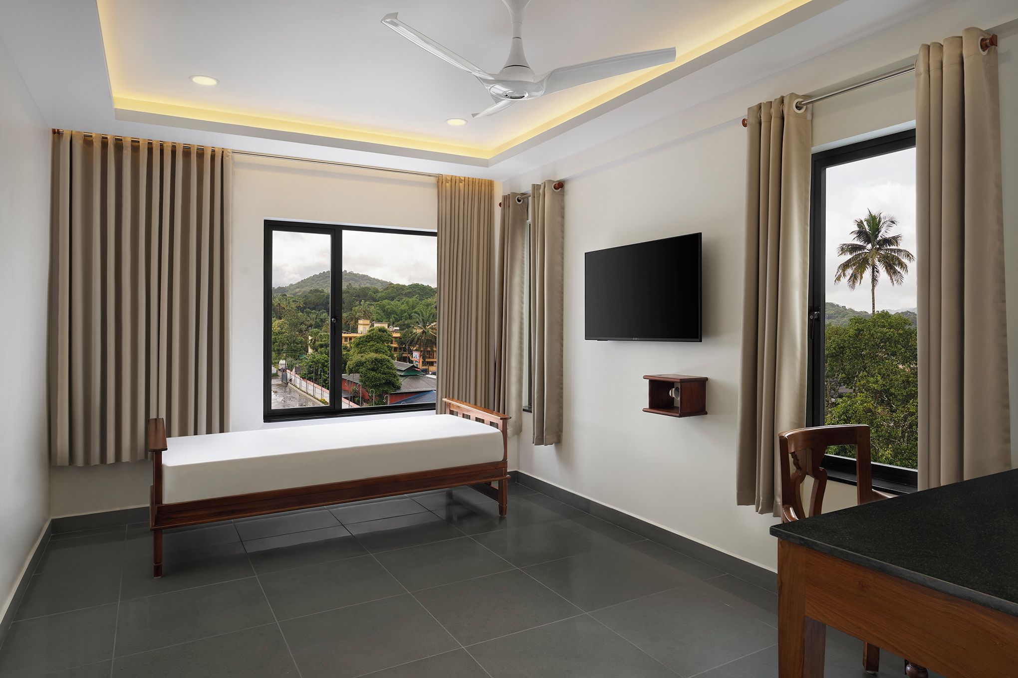 A hotel room with a bed, a TV, a window with curtains, a ceiling fan, and a view of a mountain and palm trees.