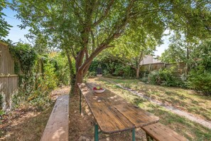 Ein Holztisch für ein Picknick mit einer Obstschale in einem Garten mit einem großen Baum, einem Holzzaun und einer Rasenfläche.