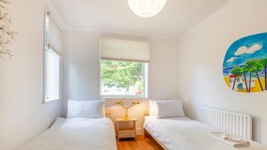 A bedroom with two single beds, a window with a view of greenery, a bedside table with a lamp, and a wall painting. - 2 Bed in Whitstable (oc-o45433) (Whitstable)