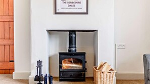 A room with a black wood-burning stove, a framed map on the wall, a basket of firewood, and a pair of boots. - 3 Bed in Cressbrook (oc-s45544) (Cressbrook)
