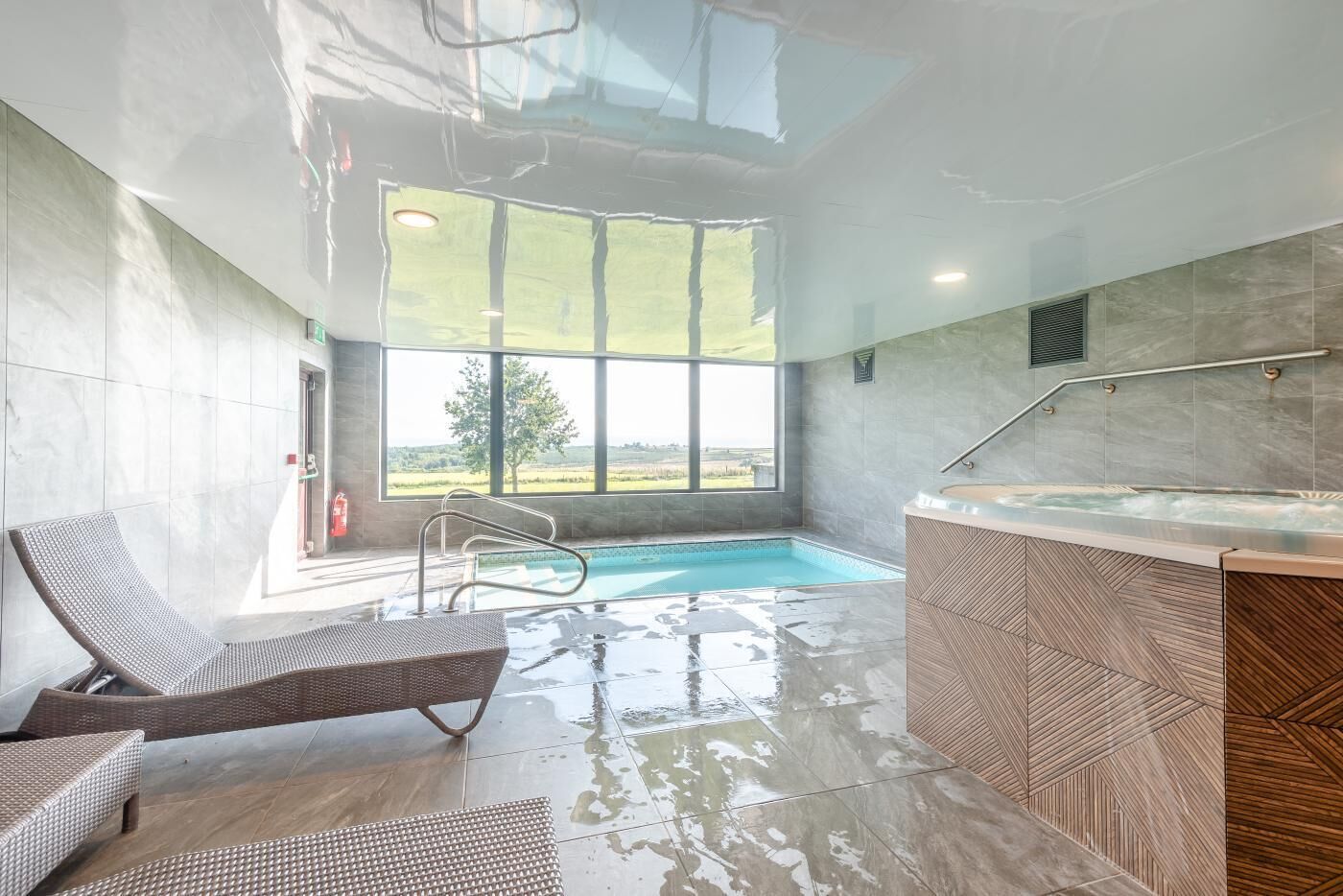 Un espace piscine intérieur moderne offrant une vue dégagée sur l’extérieur, avec un jacuzzi et des chaises longues.
