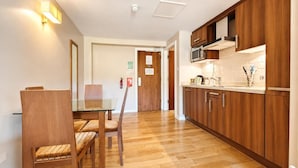A modern kitchen with wooden cabinets, a microwave, and a sink. There is a dining area with a glass table and chairs. - 1 Bed in Berrier (oc-m45743) (Hutton)