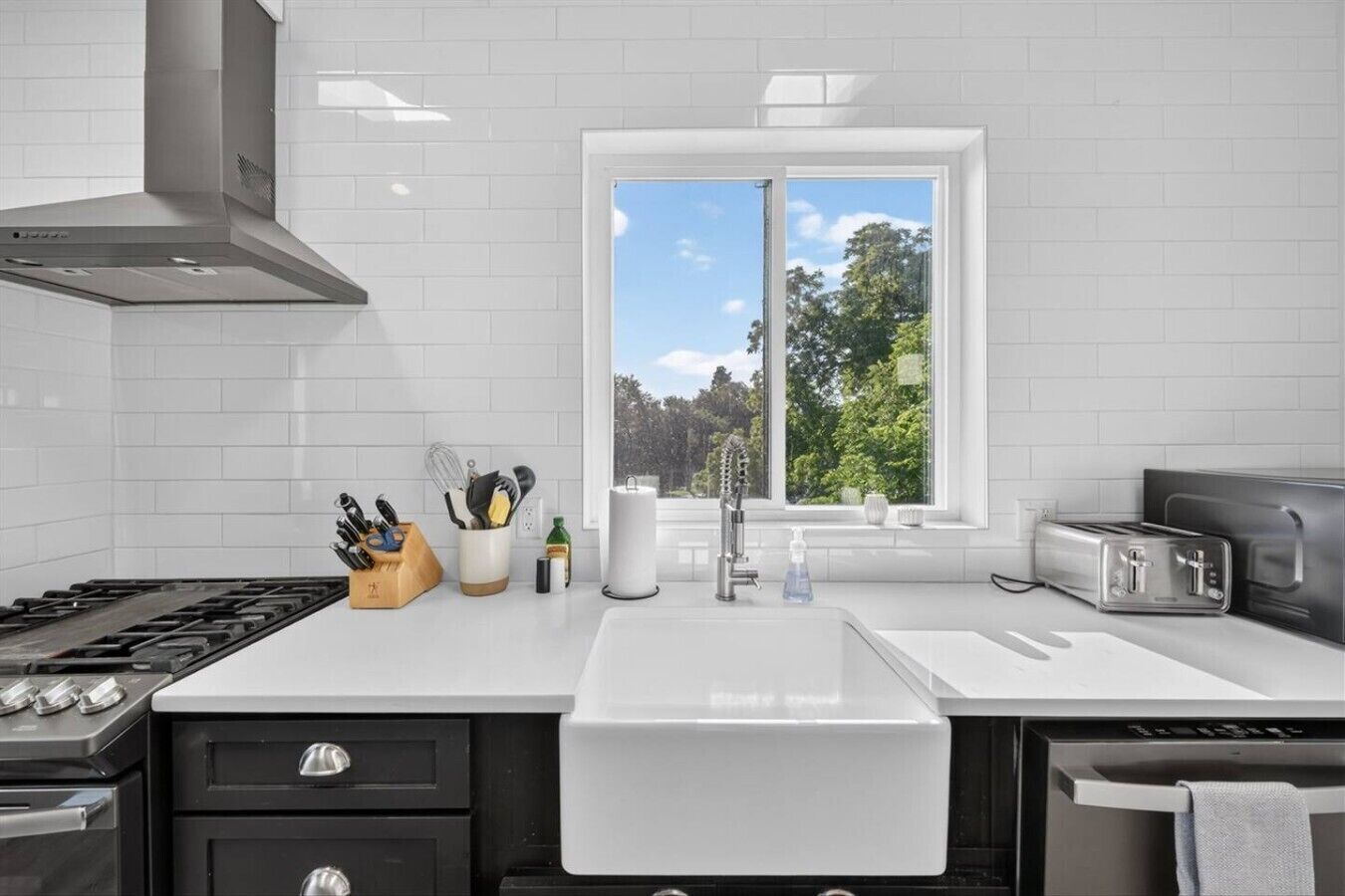 A modern kitchen with a gas stove, a stainless steel range hood, a double sink, and a window with a view of trees.