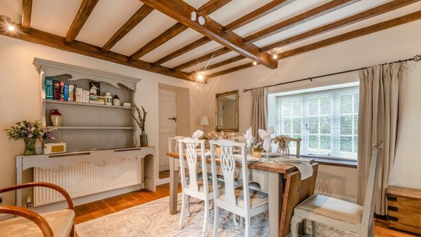 Een eethoek met een houten tafel gedekt voor vier personen, witte stoelen, een witte console met boeken en decoratieve objecten, en een raam met gordijnen.