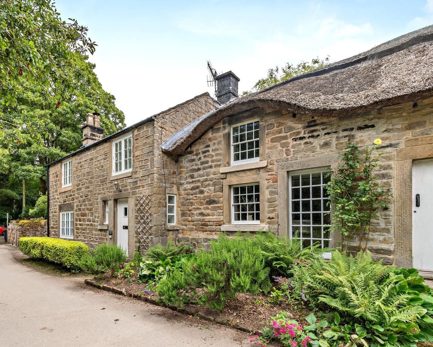 A stone building with multiple windows and a thatched roof, surrounded by lush greenery and a paved path.
