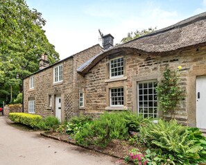 A stone building with multiple windows and a thatched roof, surrounded by lush greenery and a paved path. - 3 Bed in Baslow (oc-t44870) (Baslow)