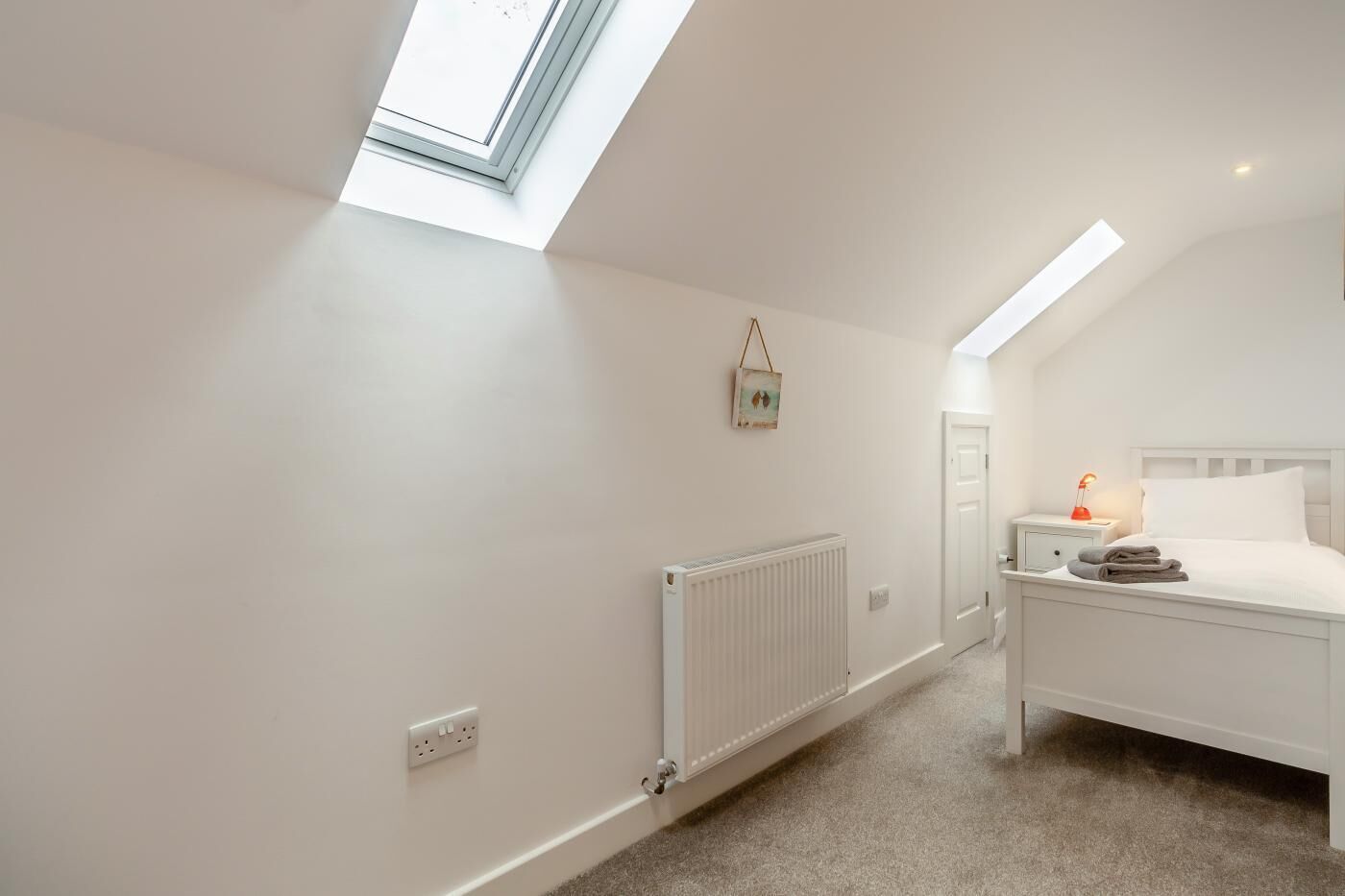 A bedroom with a skylight, a radiator, a bedside table with a lamp, and a bed with pillows.